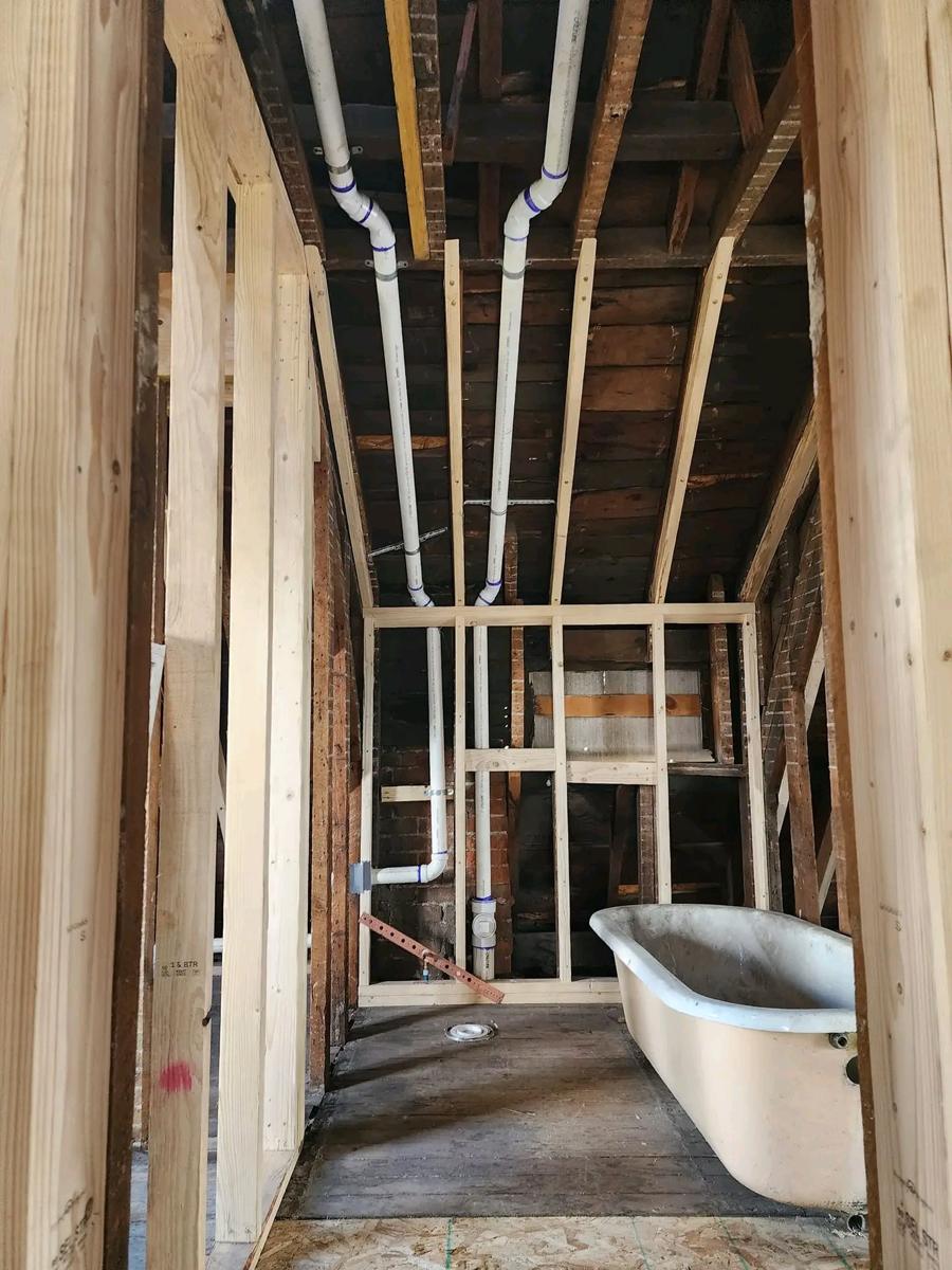 New white drain and vent pipes run along the ceiling joists toward a framed bathroom wall with a clawfoot tub waiting inside
