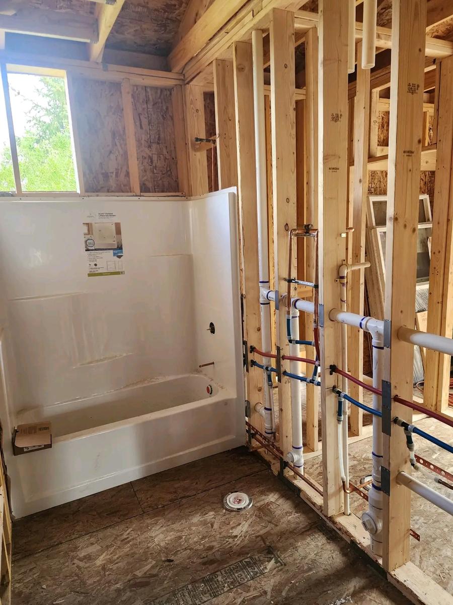 Framed bathroom with a tub and shower unit installed beside a wall of neatly run drain pipes and red and blue supply lines
